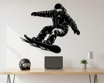May include: Black metal wall art of a snowboarder in action. The artwork depicts a person wearing a helmet and winter gear, mid-air on a snowboard. The snowboarder is set against a white wall. A black helmet, laptop, and desk lamp are on a wooden desk.