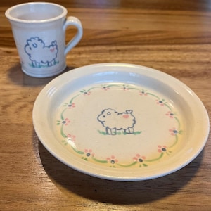May include: A ceramic mug and plate set. The off-white pieces feature a hand-drawn sheep illustration with a pink heart and blue outline. The plate has a floral border in pink, green, and yellow. The mug has a handle.