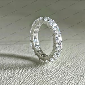 May include: A silver eternity ring featuring a continuous row of square-cut, clear gemstones. The ring is set in a polished silver band, with the gemstones closely aligned around the entire circumference. The ring is displayed on a textured, light-colored surface.