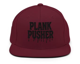 May include: A maroon baseball cap featuring the text "PLANK PUSHER" in a black, dripping font. The cap has a flat peak and a button on top. A stylish accessory for those into fitness.
