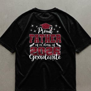 Peut inclure: T-shirt noir avec un motif rouge scintillant. Le texte indique "Proud FATHER of a class of 2026 Graduate", avec une toque de diplômé, des étoiles et des illustrations de diplômes. Le t-shirt est de couleur foncée.