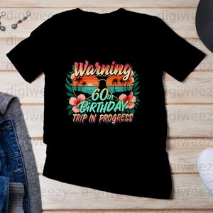 May include: Black t-shirt featuring a vibrant graphic. The design includes the text "Warning 60th Birthday Trip in Progress" with sunglasses, palm trees, and flowers. A casual wear item.
