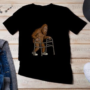 Peut inclure: T-shirt noir avec un dessin animé de Bigfoot utilisant un déambulateur. Le Bigfoot est brun et porte des baskets. Le t-shirt a un col rond et des manches courtes.