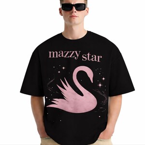 May include: Black t-shirt featuring the text "mazzy star" in pink above a pink swan graphic with stars. The shirt has short sleeves and a crew neck.