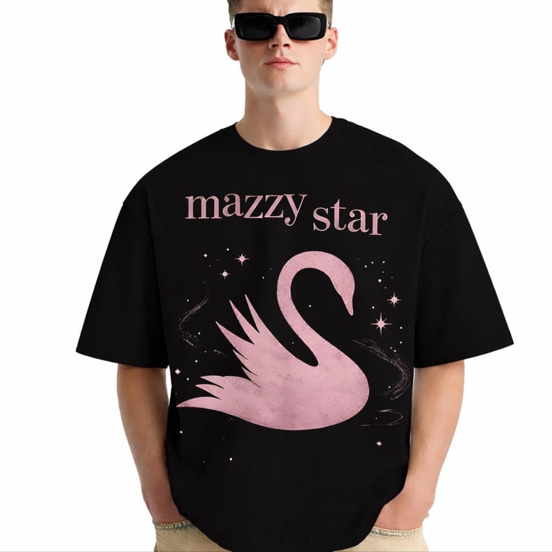 May include: Black t-shirt featuring the text "mazzy star" in pink above a pink swan graphic with stars. The shirt has short sleeves and a crew neck.