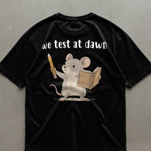 Peut inclure: T-shirt noir avec une illustration de souris de dessin animé tenant un crayon et un livre. La souris est grise et blanche, avec un livre beige et un crayon jaune. Le texte "we test at dawn" est imprimé en blanc au dos.