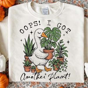 Peut inclure: Sweat-shirt crème avec un dessin d'une oie blanche tenant une plante en pot. Le texte "OOPS! I GOT ANOTHER PLANT!" est imprimé au-dessus et en dessous de l'oie. L'oie est entourée de diverses plantes en pot.