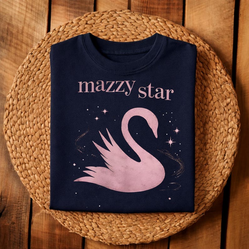 May include: Navy blue sweatshirt featuring the text "mazzy star" above a pink swan illustration with stars. The design is centered on the front of the shirt. The sweatshirt is displayed on a woven, circular placemat.