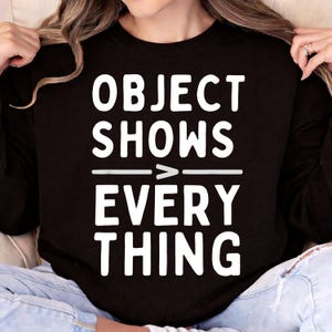 May include: Black long-sleeve sweatshirt with the white text "OBJECT SHOWS > EVERYTHING". The text is arranged in three lines with a horizontal line and arrow symbol between "SHOWS" and "EVERY".