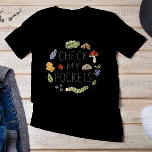 Peut inclure: T-shirt noir avec un motif graphique coloré. Le motif comprend le texte "CHECK MY POCKETS" entouré d'illustrations d'un scarabée, d'une coccinelle, d'un champignon et d'autres éléments sur le thème de la nature.
