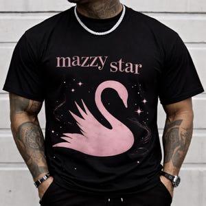 May include: Black t-shirt featuring the text "mazzy star" in pink above a pink swan graphic. The swan is set against a starry background. The shirt is a classic crew neck style.
