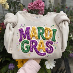 May include: Cream-colored sweatshirt with the words "MARDI GRAS" embroidered in purple, gold, and green. The letters have a textured, knitted appearance, with gold glitter accents. The sweatshirt has puffy sleeves and is held up by a person.
