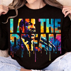 Puede incluir: Camiseta negra de manga larga con un gráfico colorido de Martin Luther King Jr. y el texto "I AM THE DREAM". El diseño utiliza tonos vibrantes de azul, amarillo, naranja y morado, con efectos de goteo de pintura. Una prenda cómoda e informal.