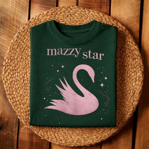 May include: Dark green t-shirt with the text "mazzy star" in pink above a pink swan illustration. The swan is surrounded by small white stars and a faint pink design. The shirt is displayed on a woven, circular mat.
