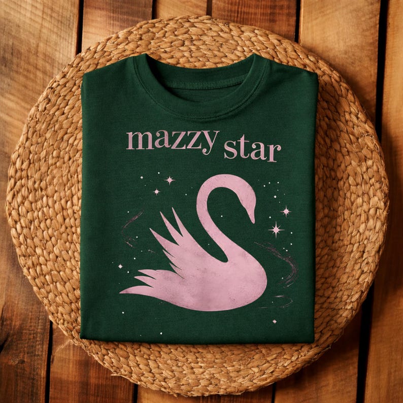 May include: Dark green t-shirt with the text "mazzy star" in pink above a pink swan illustration. The swan is surrounded by small white stars and a faint pink design. The shirt is displayed on a woven, circular mat.