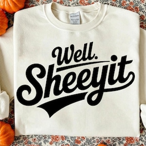 Peut inclure: Sweat-shirt crème avec l'inscription "Well. Sheeit" en lettres noires et grasses. Le texte est dans une police stylisée et vintage. Le sweat-shirt est de style col rond.