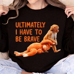 Op de afbeelding: Zwart shirt met lange mouwen met de oranje tekst "ULTIMATELY I HAVE TO BE BRAVE". Het shirt heeft een afbeelding van twee apen, waarvan één op de ander zit. De apen zijn oranje en bruin.