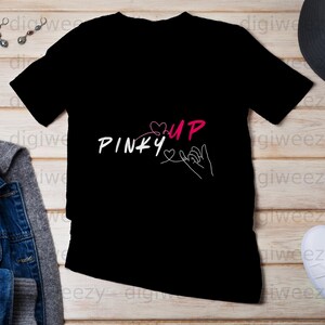 Peut inclure: T-shirt noir avec l'inscription "PINKY UP" en blanc et rose, représentant un dessin au trait de deux mains se faisant une promesse de doigts. Le motif comprend des cœurs et un trait continu.