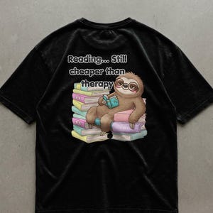 Peut inclure: T-shirt noir avec un paresseux de dessin animé portant des lunettes et lisant un livre, assis sur une pile de livres colorés. Le texte "Reading... Still cheaper than therapy" est au-dessus.