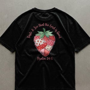 Peut inclure: T-shirt noir avec une fraise en patchwork. Le design comprend le texte "Taste & See that the Lord is Good" et "Psalm 34:8". La fraise est faite de tissus à motifs rouges, roses et blancs.