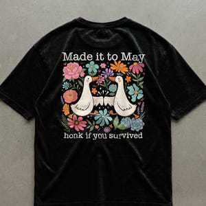 Peut inclure: T-shirt noir avec un motif floral et deux oies blanches. Le texte "Made it to May" est au-dessus du motif, et "honk if you survived" en dessous. Le motif comprend des fleurs roses, oranges, bleues et violettes.