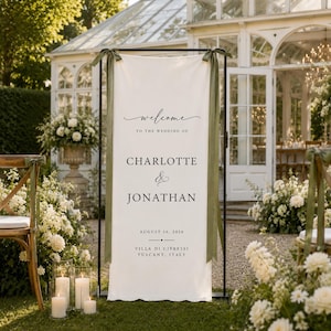Op de afbeelding: Een verticaal trouwbord van gebroken wit textiel hangend aan een zwart metalen frame, met zwarte tekst: Welcome to the wedding of Charlotte & Jonathan, August 16, 2026, Villa di Cipressi, Tuscany, Italy, afgewerkt met olijfgroene linten.