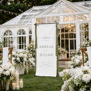 May include: A tall, vertical white wedding welcome sign featuring black text that reads Welcome to the wedding of Amelia and Julian, June 29, 2024, Napa, California. The sign is suspended from a black metal frame with olive green ribbons.