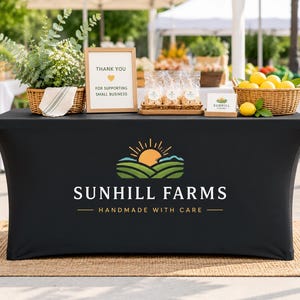 Könnte beinhalten: Eine schwarze, passgenaue Tischdecke mit dem Sunhill Farms-Logo, das einen Sonnenaufgang über grünen Hügeln und den Text "Handmade with Care" zeigt. Auf dem Tisch stehen ein Weidenkorb mit Grünpflanzen, ein "Thank You"-Schild und Zitronen.