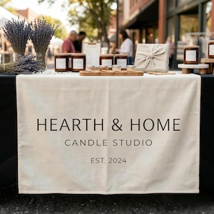 May include: A display table at an outdoor market features candles, lavender, and a wrapped gift. The table is draped with a cloth printed with "HEARTH & HOME CANDLE STUDIO EST. 2024". Several candles with wooden lids are arranged on the table.