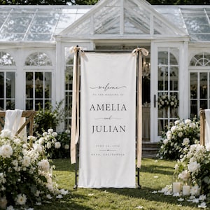 May include: A vertical white fabric wedding welcome sign suspended on a black metal frame with beige ribbons. The text reads: Welcome to the wedding of Amelia and Julian, June 20, 2026, Napa, California, in black serif and script typography.