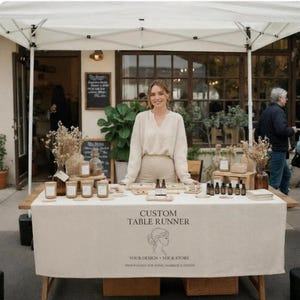 Puede incluir: Una mujer está detrás de una mesa en un mercado al aire libre, exhibiendo productos. La mesa está cubierta con un camino de mesa beige personalizado con texto y un logotipo. Varios artículos, incluyendo velas y aceites esenciales, están expuestos.