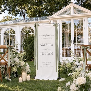 May include: A tall, rectangular off-white fabric wedding welcome sign suspended on a black metal frame. The sign features black text reading "Welcome to the wedding of Amelia and Julian, June 20, 2024, Napa, California" with sage green ribbons.