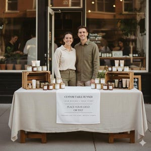 May include: A couple stands behind a table displaying candles and skincare products. The table is covered with a beige cloth and has a banner that reads "CUSTOM TABLE RUNNER". The products are arranged on wooden crates and the table. The background is a storefront.
