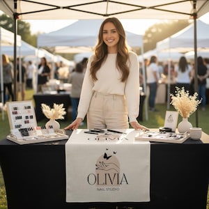 May include: A woman at an outdoor market stall with a table displaying nail products. The table has a black cloth and a cream runner with "OLIVIA NAIL STUDIO". Nail polish samples and other items are visible.
