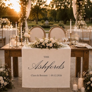 Puede incluir: Un camino de mesa de tela blanca sobre una mesa de madera, con el texto impreso THE Ashfords Clara & Bennett | 09.12.2026. El camino está decorado con un arreglo floral central de rosas blancas y follaje, flanqueado por velas altas.