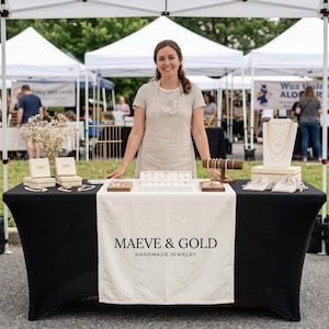 May include: A black table display featuring a white runner with the text MAEVE & GOLD HANDMADE JEWELRY. The table holds jewellery boxes, a necklace display stand, a bracelet holder, and various jewellery pieces arranged on white trays and small pouches.