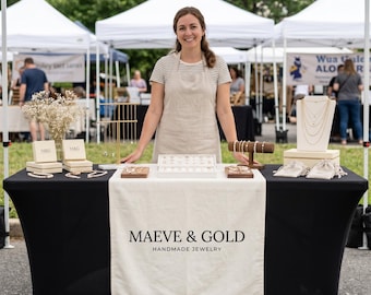 Puede incluir: Una mesa negra con un camino de mesa blanco que muestra el texto MAEVE & GOLD HANDMADE JEWELRY. La mesa contiene cajas de joyería, un expositor de collares, un soporte para pulseras y varias piezas de joyería sobre bandejas y bolsitas.