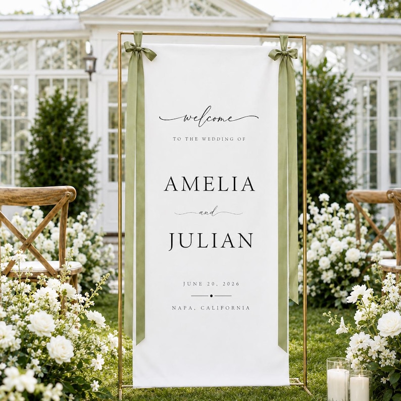 May include: A tall white rectangular wedding welcome sign suspended on a gold metal frame. The sign features black text reading Welcome to the wedding of Amelia and Julian, June 20, 2026, Napa, California. Two sage green ribbons are tied at the top.