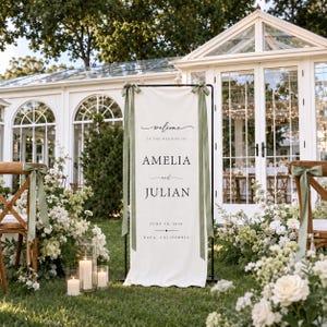 May include: A tall, rectangular off-white fabric wedding welcome sign suspended on a black metal frame. The sign features black text reading "Welcome to the wedding of Amelia and Julian, June 20, 2024, Napa, California" with sage green ribbons.