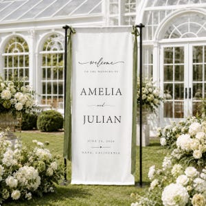 May include: A tall white vertical fabric wedding welcome sign suspended from a black metal stand with olive green ribbons. The text reads: Welcome to the wedding of Amelia and Julian, June 20, 2026, Napa, California, in black serif and script fonts.