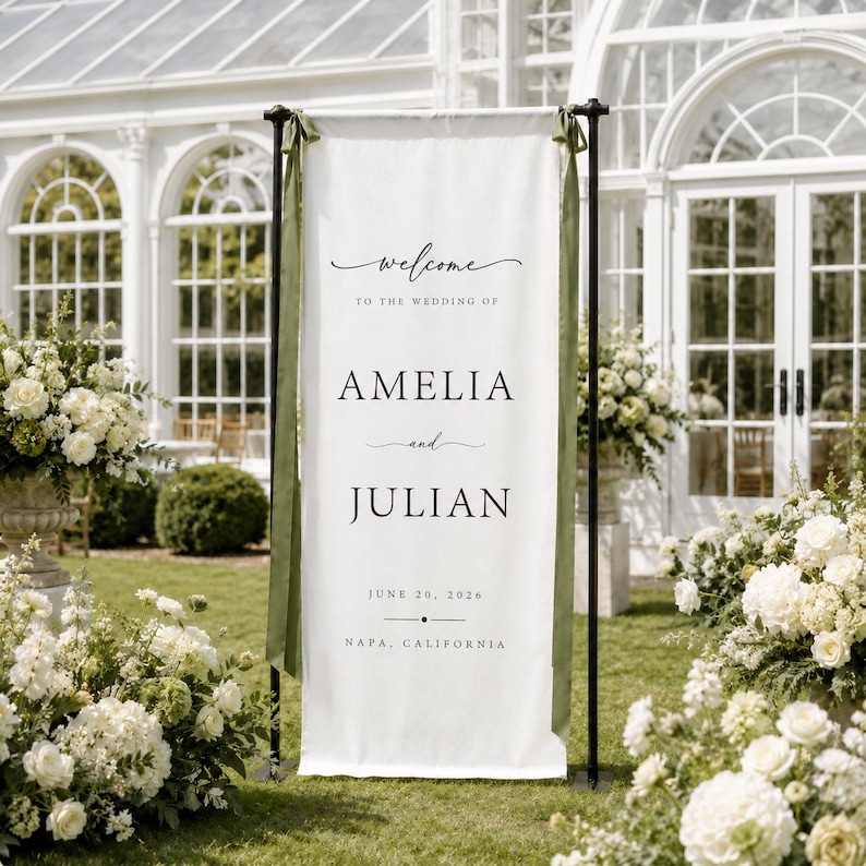 May include: A tall white vertical fabric wedding welcome sign suspended from a black metal stand with olive green ribbons. The text reads: Welcome to the wedding of Amelia and Julian, June 20, 2026, Napa, California, in black serif and script fonts.