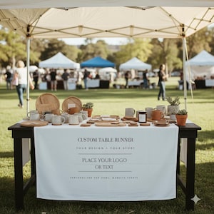 Può includere: Un'esposizione di mercato all'aperto con un tavolo nero coperto da un runner personalizzato bianco. Il tavolo mostra varie tazze in ceramica, vassoi di legno, sottobicchieri e piccole piante in vaso. Lo sfondo mostra altre bancarelle e persone.
