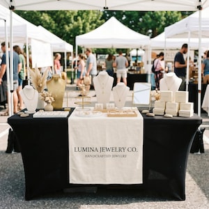 May include: A black table display for Lumina Jewelry Co. featuring a cream-colored runner with the text Lumina Jewelry Co. Handcrafted Jewelry. The setup includes multiple jewelry busts, gold-toned earring stands, a small mirror, and stacked boxes.