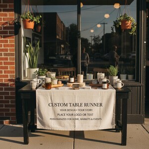 May include: A black table with a white table runner displaying the text "CUSTOM TABLE RUNNER". The table is set with mugs, candles, and plants. The scene is in front of a storefront with hanging plants.