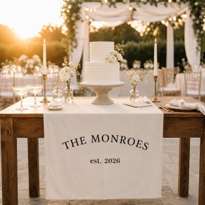 Op de afbeelding: Een witte bruidstaart met twee lagen op een voetstuk staat op een houten tafel met een crèmekleurige tafelloper. De loper bevat de zwarte tekst THE MONROES en est. 2026. Twee hoge witte kaarsen in gouden houders staan naast de taart.