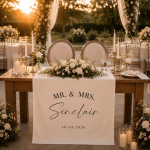 May include: A white fabric table runner draped over a wooden table, featuring the black printed text MR. & MRS. SINCLAIR and the date 10.03.2026. The runner is decorated with a centrepiece of white roses and daisies, flanked by tall candles.