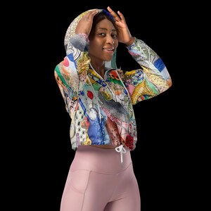 May include: A lightweight, cropped windbreaker jacket featuring a vibrant, multi-colored abstract graphic print. The jacket includes a hood with adjustable white drawstrings, long sleeves, and a front zipper closure, paired with solid light pink leggings.
