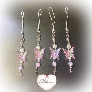 Op de afbeelding: Vier telefoonbedels met iriserende roze vlinderhangers, witte hartvormige kralen, kleine zilveren sterbedels en ronde pastelkleurige kralen aan zilverkleurige metalen kettinkjes met witte lusjes. Een hartvormig logo met de tekst Milamore staat onderaan.