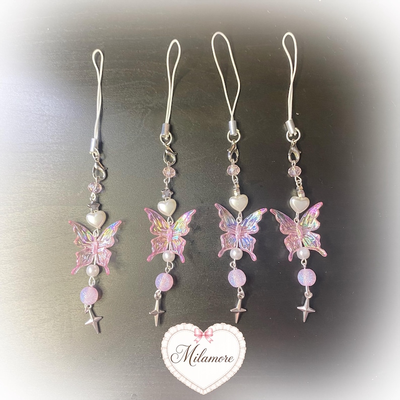 May include: Four phone charms featuring iridescent pink butterfly pendants, white heart beads, small silver star charms, and round pastel beads on silver-toned metal chains with white loops. A heart-shaped logo with the text Milamore is at the bottom.