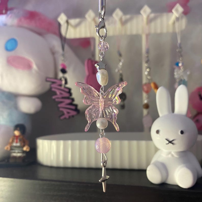 May include: A phone charm featuring a translucent iridescent pink butterfly bead as the centerpiece. The charm includes a silver-toned lobster clasp, a small silver star charm, a white pearl-like bead, a light pink glitter bead, and a silver cross charm.